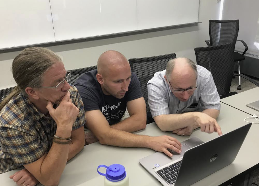 Three men sit at a desk and consider what's presented on a laptop computer while one of them gestures toward it